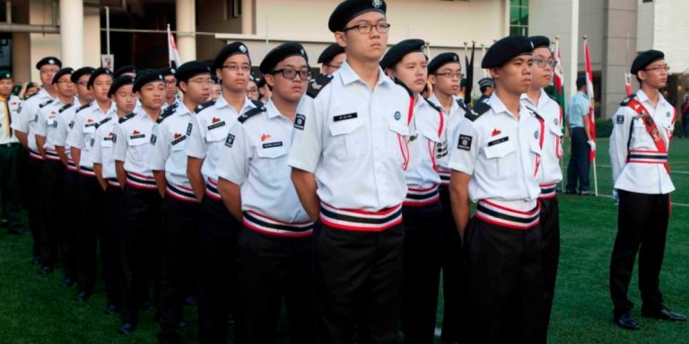 ACS(I) St John's Brigade members in white uniforms march in formation during a parade or drill competition, with red accents visible on their uniforms.
